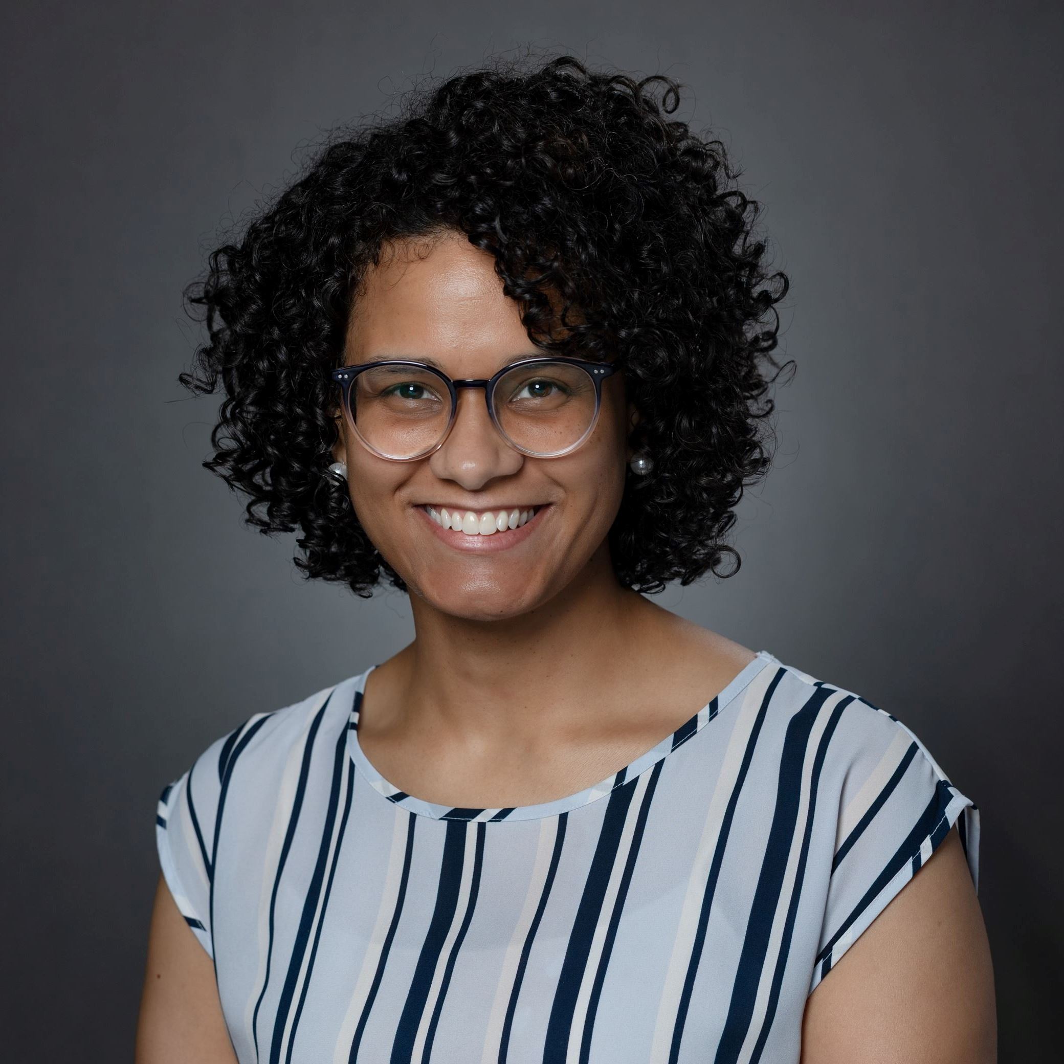 Headshot of Ashlee Smith female with short curly dark hair and glasses wearing pinstripe blouse